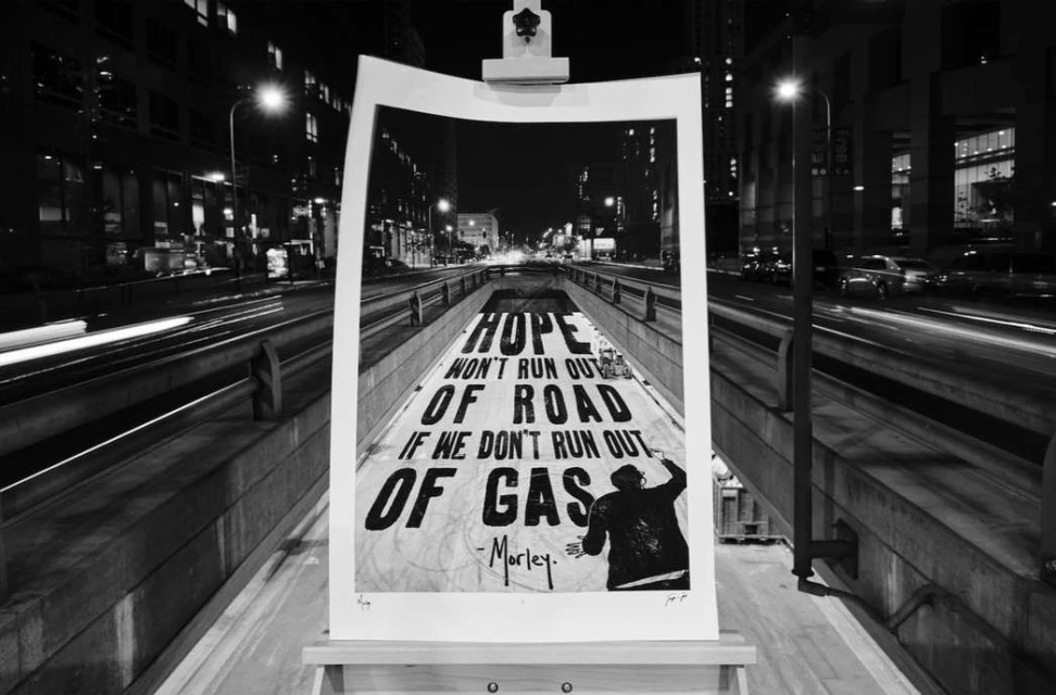 Black and white photograph showcasing street art on an easel with an urban city street and nighttime light trails in the background.