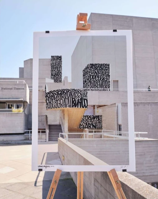 Architectural building art installation with typography on white panels supported by a wooden easel in front of a Brutalist structure.