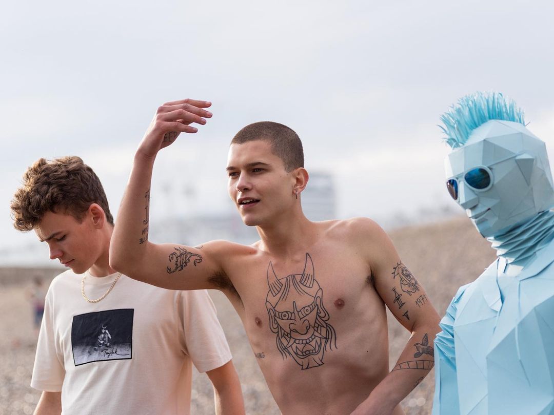 Three people stand on a beach: one with body tattoos, another in a graphic tee, and another in a geometric blue full-body suit.