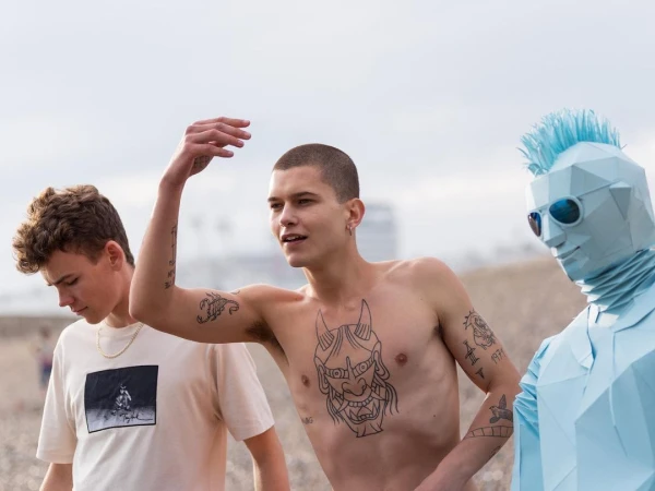 Three people stand on a beach: one with body tattoos, another in a graphic tee, and another in a geometric blue full-body suit.