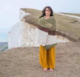 Woman standing on a cliff edge wrapped in a striped blanket and wearing yellow trousers, photographed by Joseph Ford.