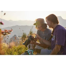 Two people seated outdoors in a mountainous landscape, reviewing images on a digital camera together.