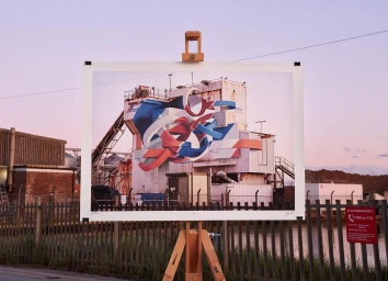 A photograph on an easel set up outdoors showing a painted industrial building with matching ribbons blending seamlessly into the scene.