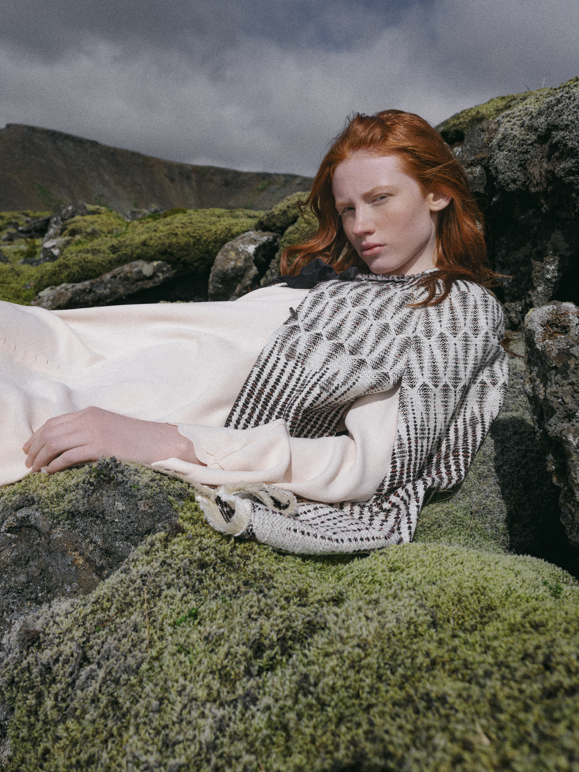 Red-haired woman in patterned sweater lies on mossy rocks in a natural outdoor setting, with mountains and cloudy sky in background.