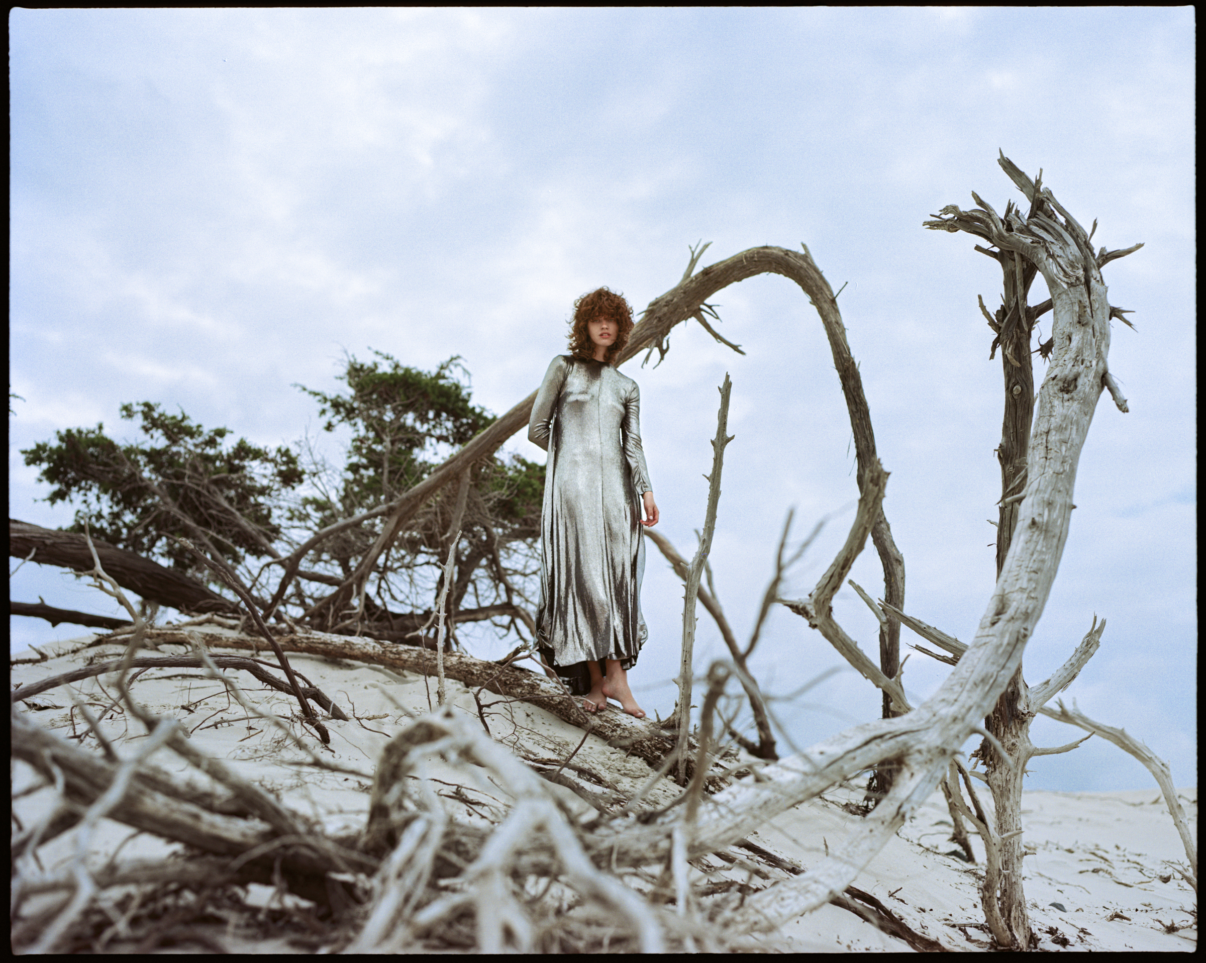 Woman in a shiny silver dress stands on a sandy dune surrounded by driftwood against a cloudy sky in a fashion photoshoot.