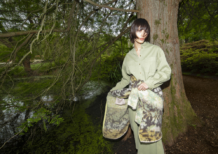 An adult female poses in a forest near water, wearing a green outfit with a patterned scarf, next to a thick tree trunk in daylight.