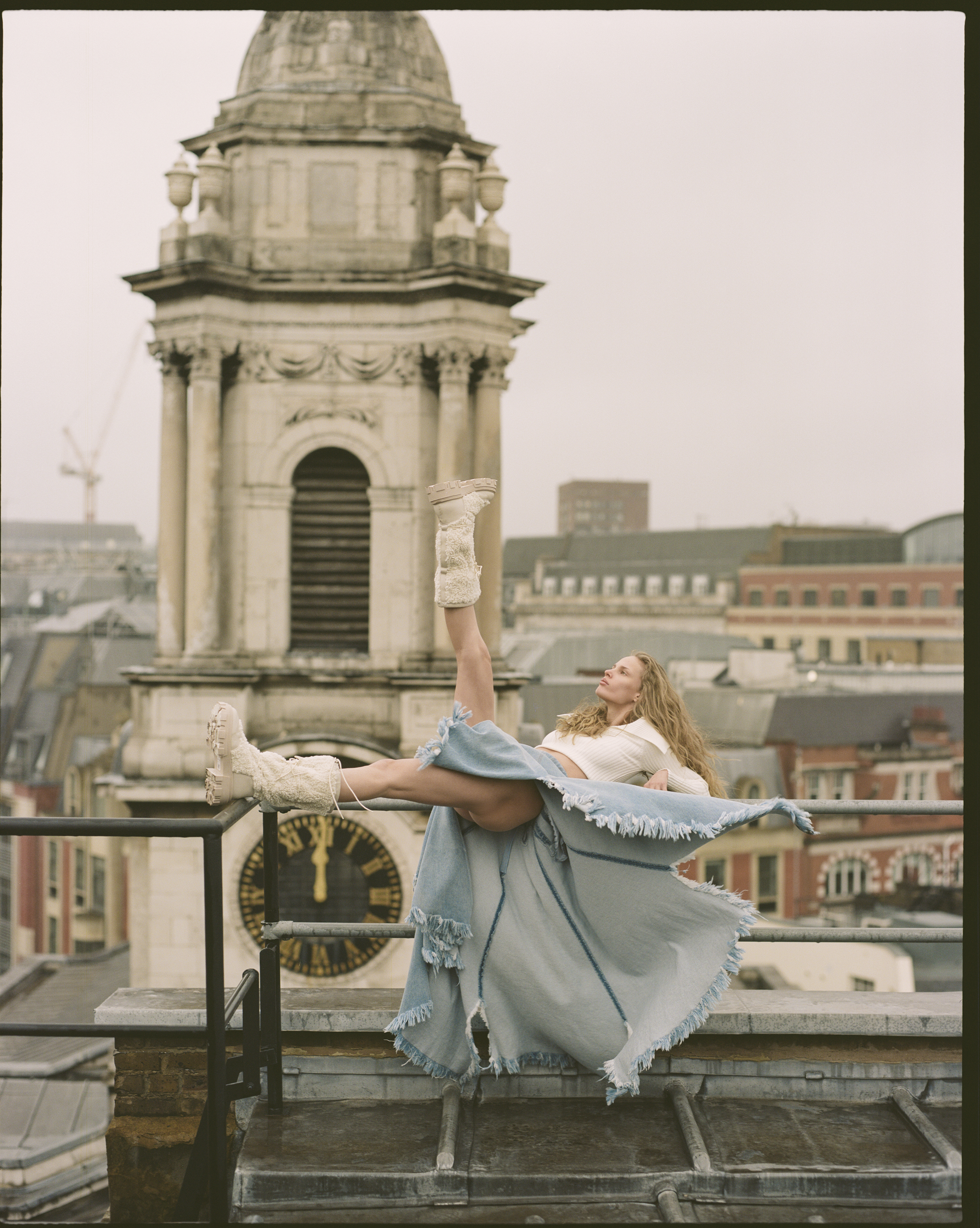 Adult female fashion model in a long blue skirt and off-white boots poses on a rooftop with a building tower in the background.