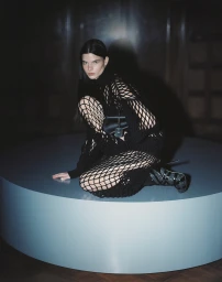 Woman in black mesh outfit crouching on a round teal platform holding a dark handbag