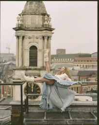Model reclining on a rooftop railing wearing a draped frayed denim skirt and chunky cream boots with a historic clock tower behind.