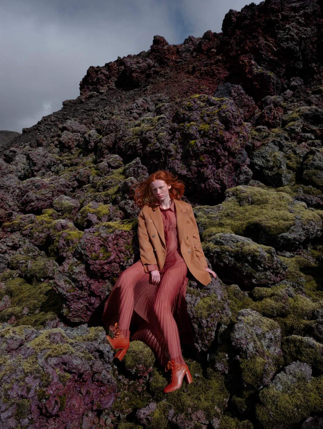Red-haired model in sheer pleated dress and tan blazer sitting on moss-covered volcanic rocks