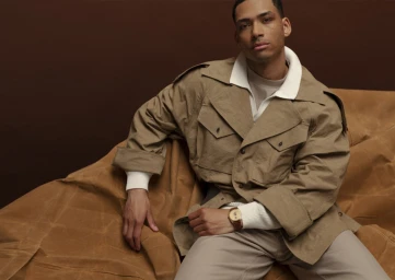 Man wearing a beige jacket and light trousers seated against a brown background, photographed by Zuzanna Blur