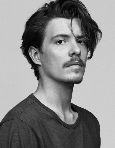Monochrome portrait of a young adult male with wavy dark hair and a mustache, wearing a casual T-shirt against a plain background.