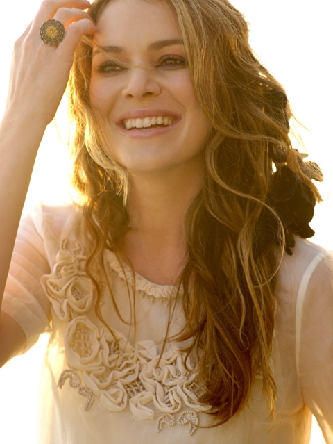 Portrait of a fair-skinned adult female with long brown hair, wearing a sheer floral-embroidered blouse, and a gold ring on her finger.
