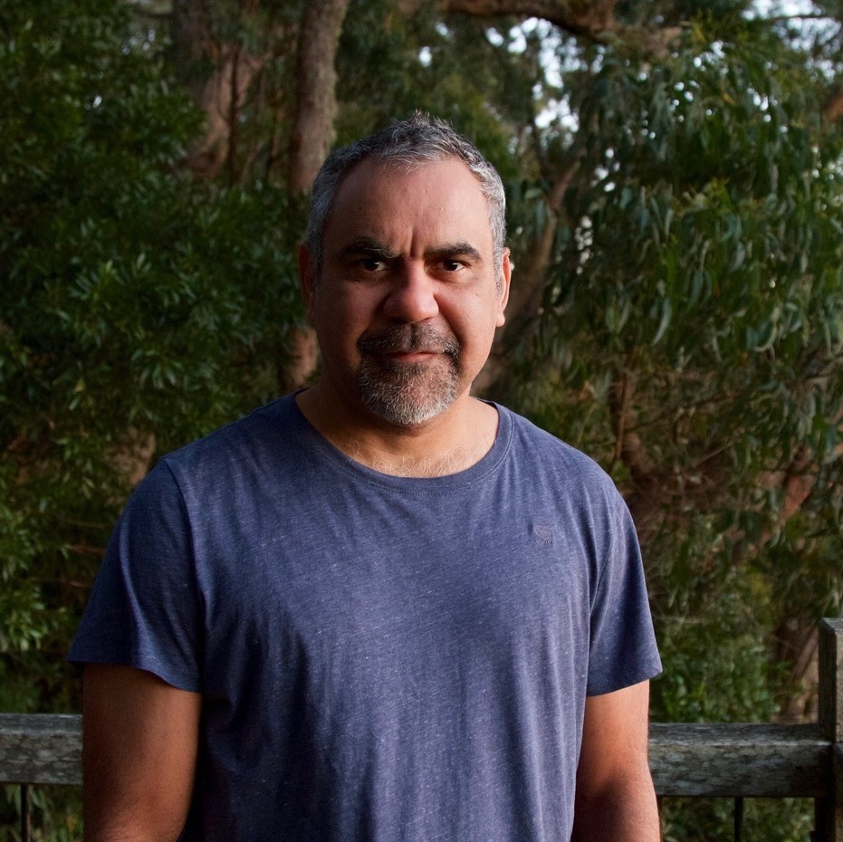 Portrait of a middle-aged man with salt and pepper hair, a beard, and a blue t-shirt, standing outdoors against a green, leafy backdrop.