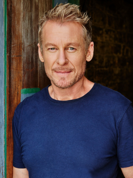 Headshot of a blonde adult male with blue eyes wearing a navy t-shirt, posing in front of a wooden wall with blue and green accents.