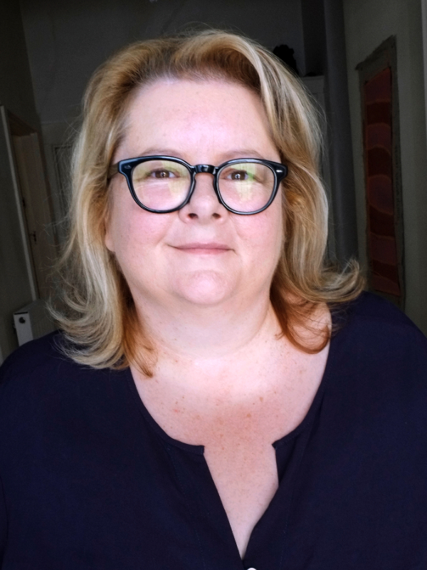 Portrait of an adult woman with blonde hair and glasses wearing a dark blue top in an indoor setting with natural lighting.