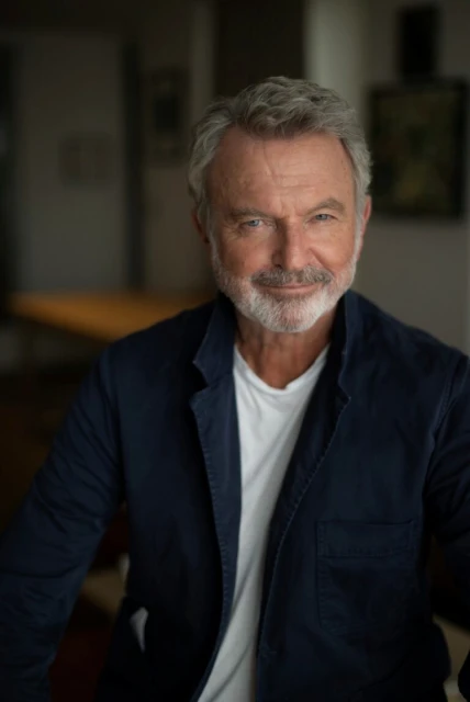 Portrait of Sam Neill wearing a dark blazer and white t-shirt indoors with soft natural lighting and blurred background.