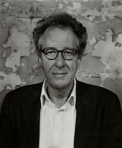Monochrome portrait of an adult male wearing glasses and a dark jacket, posing in front of a textured wall with a neutral color palette.