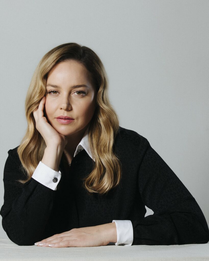 Portrait of an adult woman with blonde hair wearing a black sweater and white collared shirt, resting her chin on her hand.