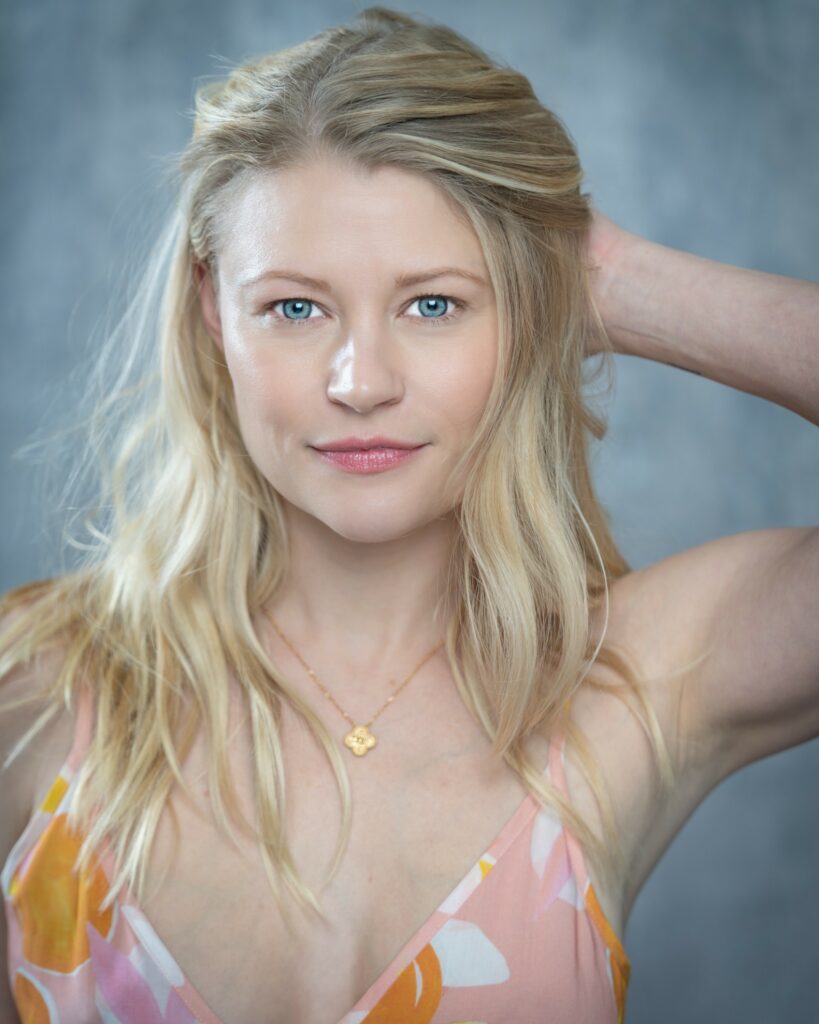 Portrait of a blonde woman with blue eyes, wearing a patterned top and gold necklace, posing in front of a textured gray background.