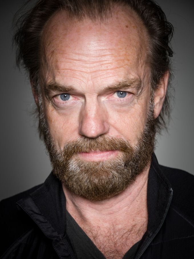 Portrait of a mature adult male with a beard looking at the camera, wearing a black jacket, against a grey studio backdrop.