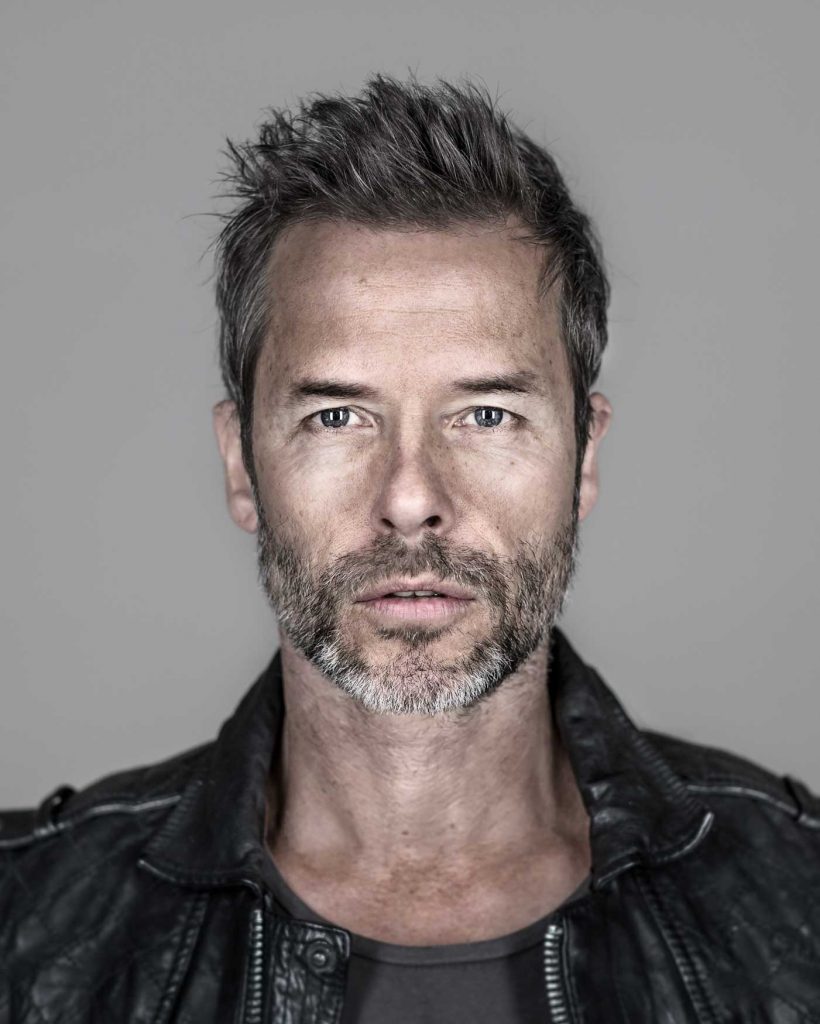 Portrait of a mature adult male with graying hair and a beard, wearing a black leather jacket against a neutral gray backdrop.