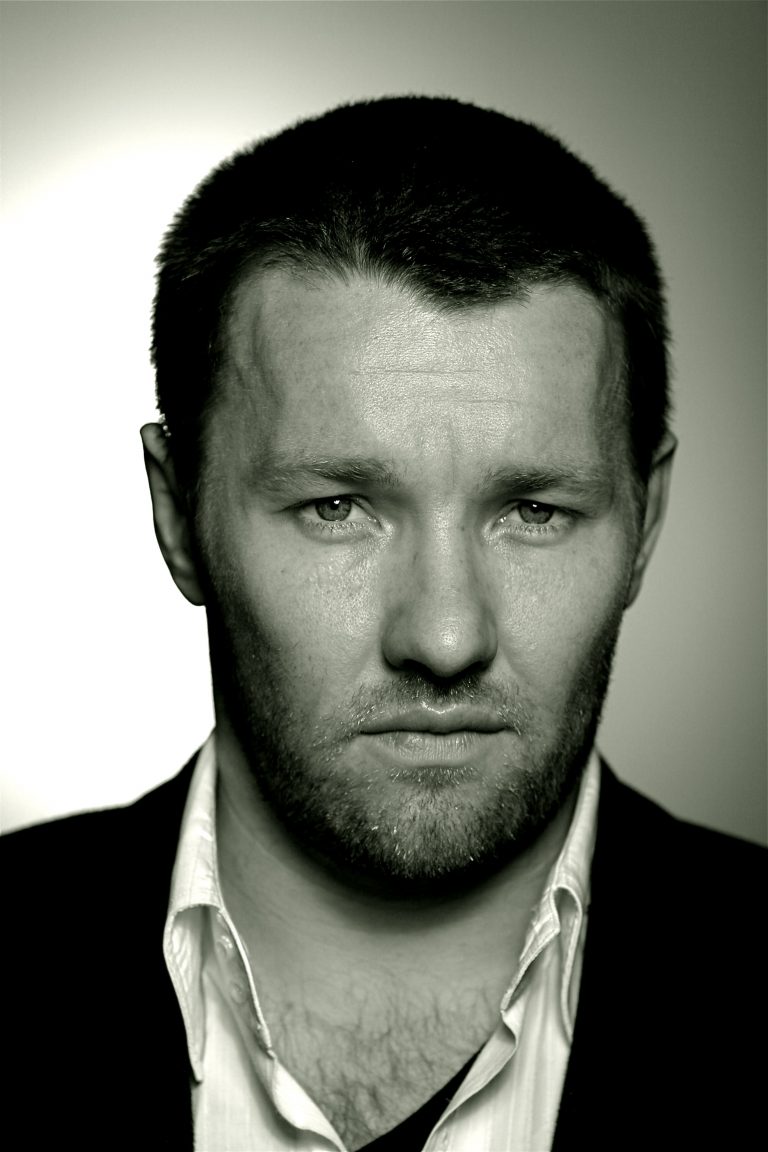 Close-up portrait of Joel Edgerton with short hair and facial hair, wearing a white shirt and dark jacket against a light gradient background.