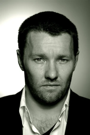 Close-up portrait of Joel Edgerton with short hair and facial hair, wearing a white shirt and dark jacket against a light gradient background.