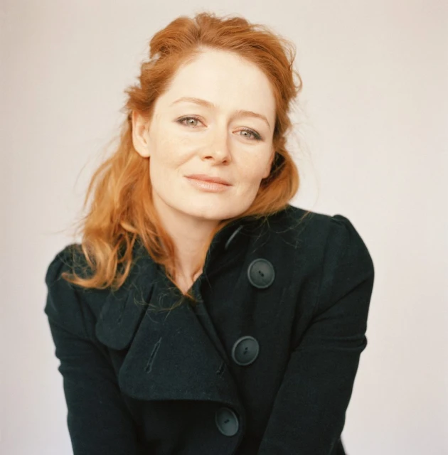 Red-haired woman in a dark buttoned coat, posing in a studio portrait. Close-up shot of a stylish, casually dressed woman.