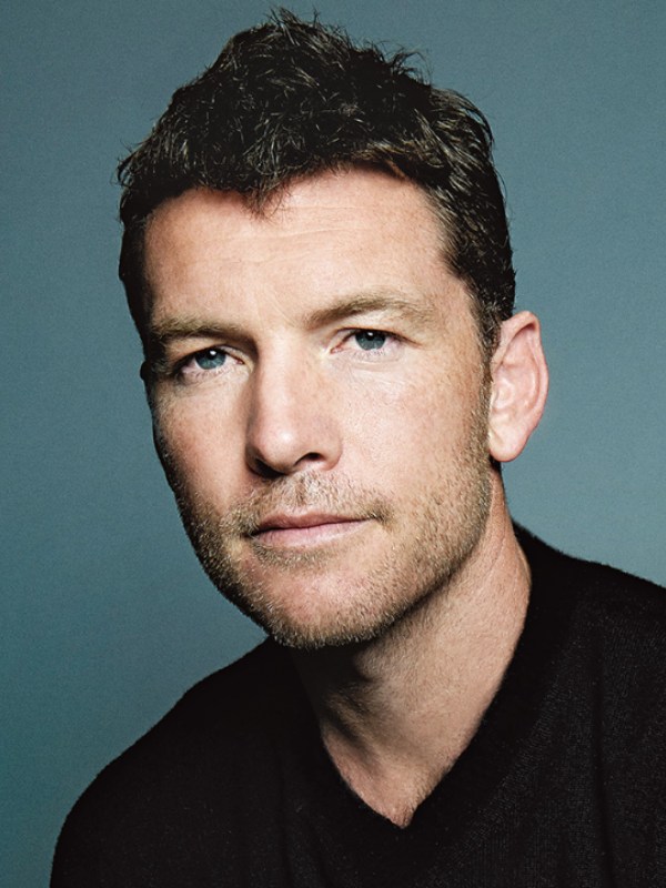 Portrait of an adult male with short dark hair and a light beard, wearing a black top against a blue-grey backdrop in a studio setting.