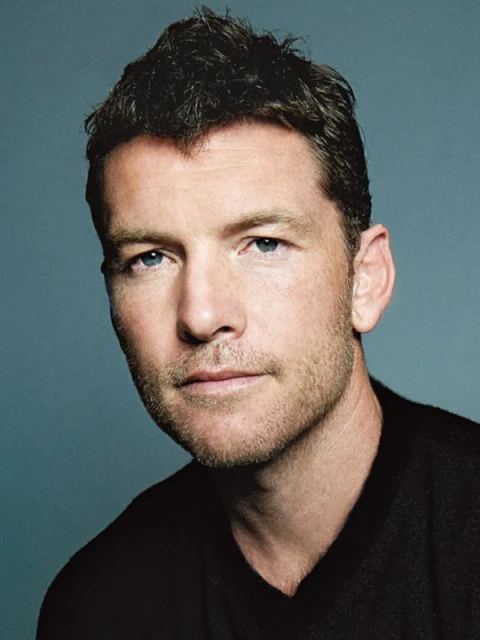 Portrait of an adult male with short dark hair and a light beard, wearing a black top against a blue-grey backdrop in a studio setting.