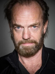 Portrait of Hugo Weaving with a grey-streaked beard and blue eyes wearing a black jacket against a grey background.