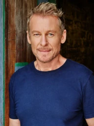 Portrait of a middle-aged man with short blonde hair wearing a dark blue t-shirt standing against a textured wooden background.