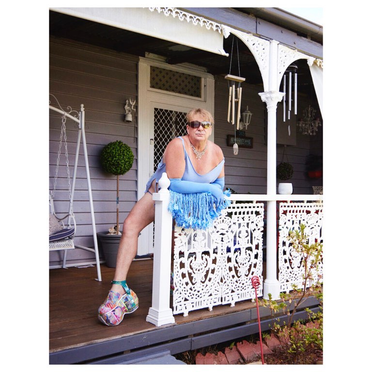 Person on porch in blue dress, matching gloves and statement shoes. The house is grey with white trim and lush green plants.
