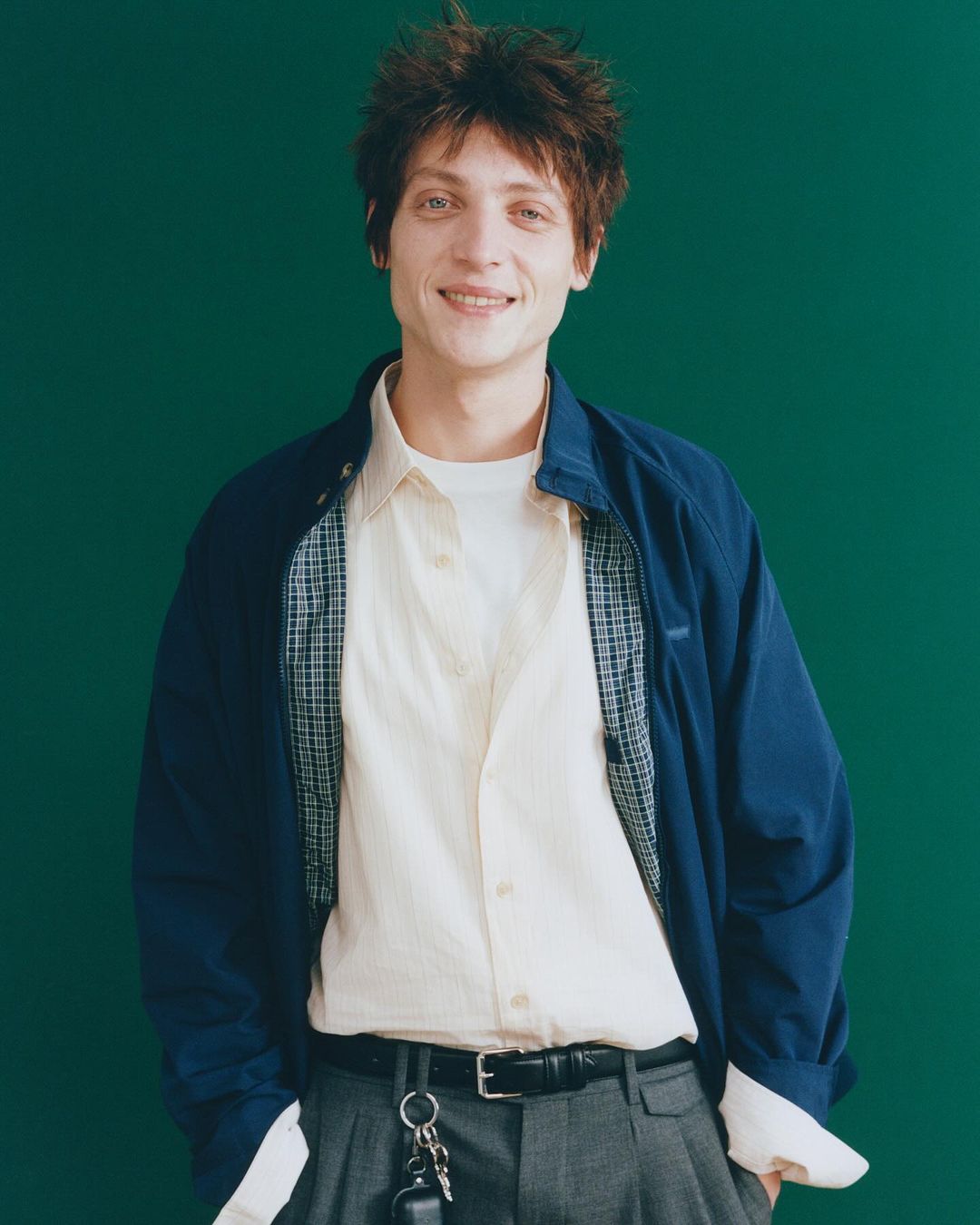 Portrait of a young adult man in a blue jacket and grey trousers against a green background, smiling with messy brown hair.