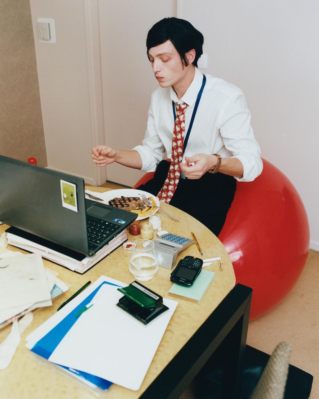Man sits on a red exercise ball at a desk, wearing a shirt and tie, using a laptop with food and office supplies scattered around the desk.