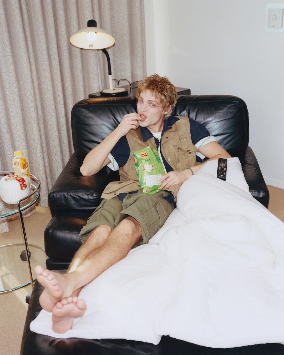 Young adult male lounges on a leather chaise, eating chips under a lamp in an indoor setting, covered with a white blanket.