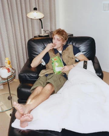 Young adult male lounges on a leather chaise, eating chips under a lamp in an indoor setting, covered with a white blanket.