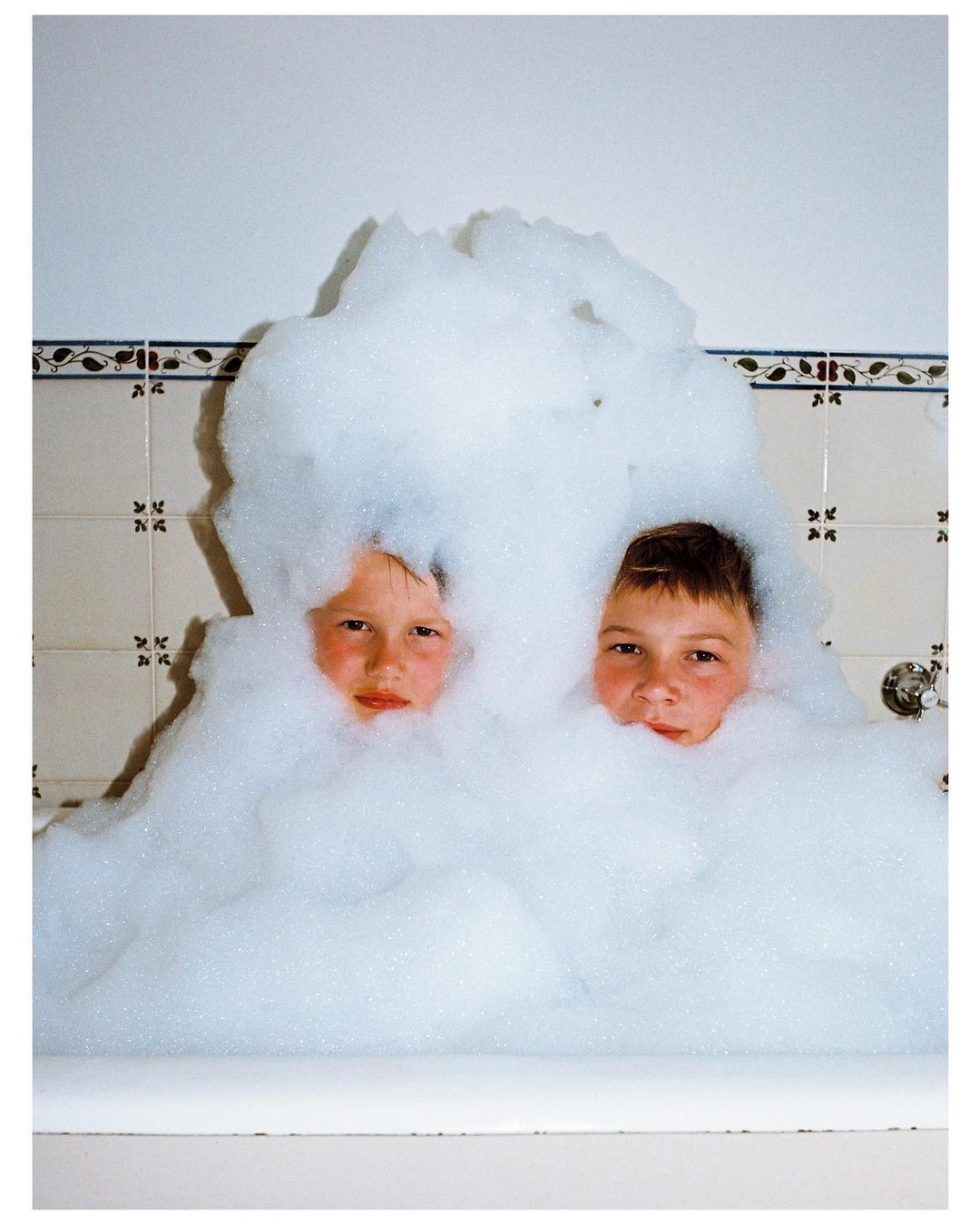 Two children are partially submerged in a bathtub filled with large amounts of white soap bubbles, faces visible, bathroom tiles in the background.