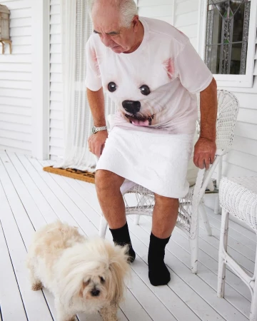 Elderly man wearing a dog-print t-shirt and white towel stands up from a white wicker chair with a small fluffy dog on a porch.