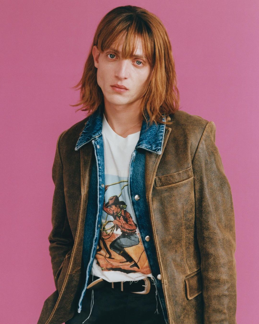 Portrait of a fair-skinned young adult with long brown hair wearing a brown jacket and cowboy graphic t-shirt against a pink background.