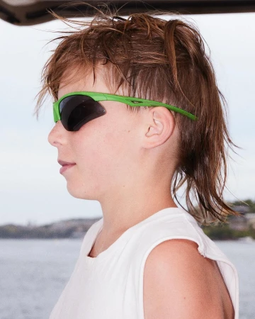 Profile portrait of a child wearing bright green wraparound sports sunglasses and a white sleeveless top against a bright background.