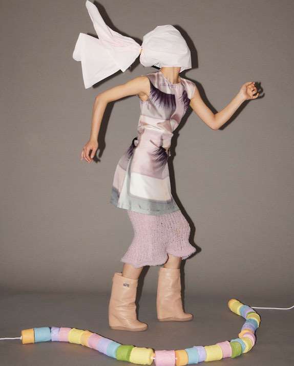 Woman in artistic fashion ensemble with head covering and neutral tones striking a playful pose against a grey backdrop, a colorful beaded string.