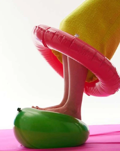 Low angle view of legs and feet on a green balloon under an inflatable red ring, set on a pink mat against a white background.