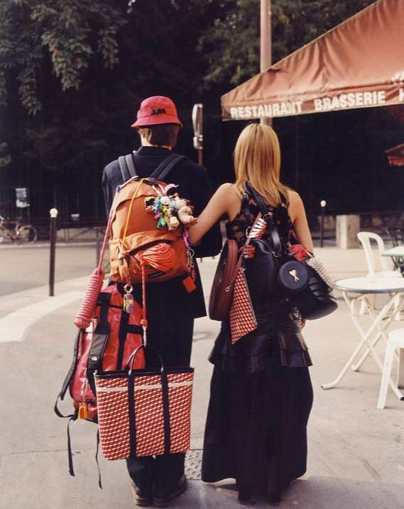 Back view of a stylish couple with overloaded backpacks in an urban environment next to a restaurant with warm lighting and vintage tones.