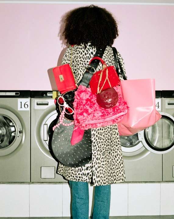 A person with curly hair is seen from behind wearing a leopard print coat, blue jeans, and carrying red and pink bags, standing in a laundromat.