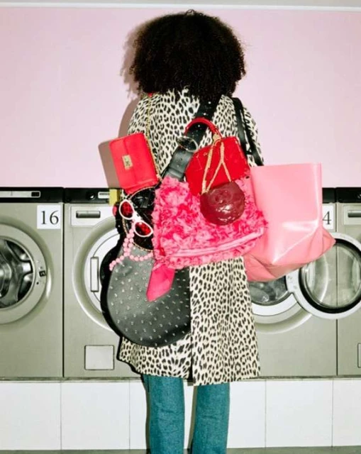 A person with curly hair is seen from behind wearing a leopard print coat, blue jeans, and carrying red and pink bags, standing in a laundromat.