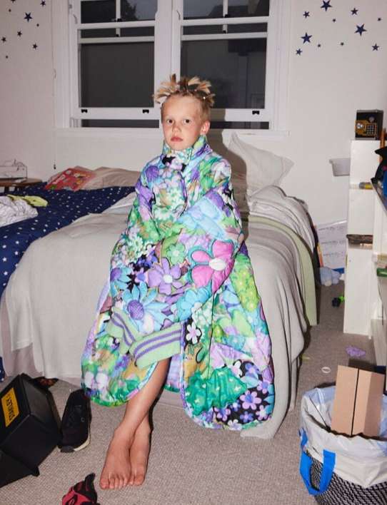 Child seated on a bed, wrapped in a floral quilted blanket, looking directly at the camera in a bedroom setting with natural light.