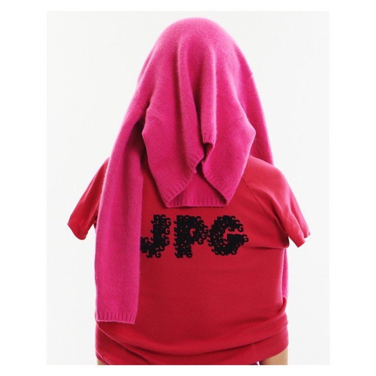 Person wearing red shirt with JPG text covered in pink sweater in a white indoor setting. Casual style. Fashion photography.