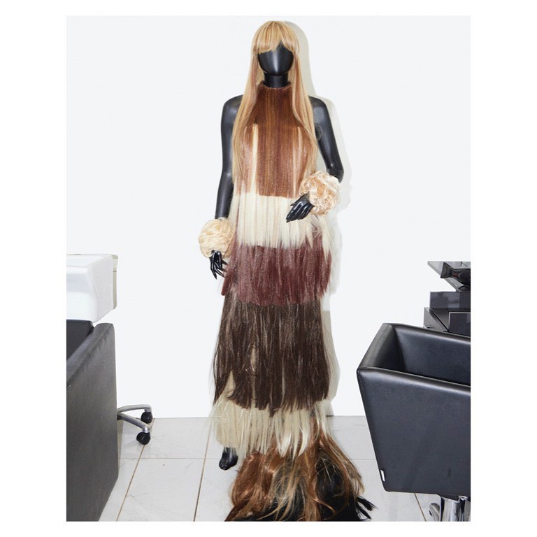 Mannequin dressed in a tiered gown made from various shades of human hair, accessorized with large hair-covered bracelets, against white backdrop.
